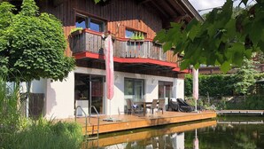 A two-story wooden house with a balcony, a dining area with chairs and a table, and a small dock extending into a pond. - Room-charming Guest Room in Ebbs Nature (Ebbs)