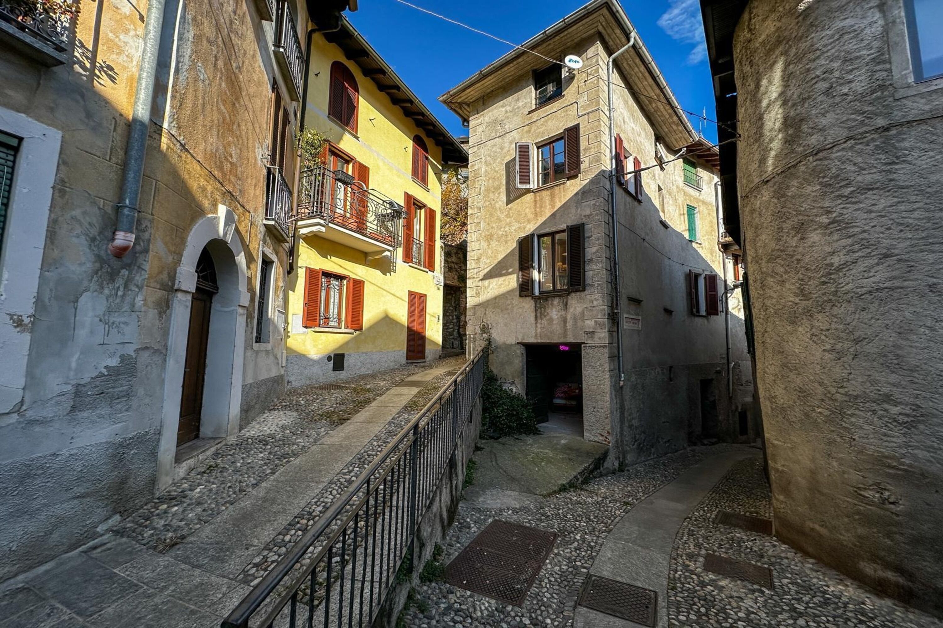 A narrow alleyway between buildings with a metal railing, stone walls, and a clear blue sky.