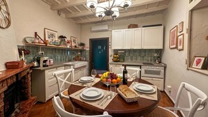 A dining area with a wooden table set for four, white chairs, a chandelier, and a kitchen in the background. - La Fetta Di Cima, Porlezza, Italy (Porlezza)