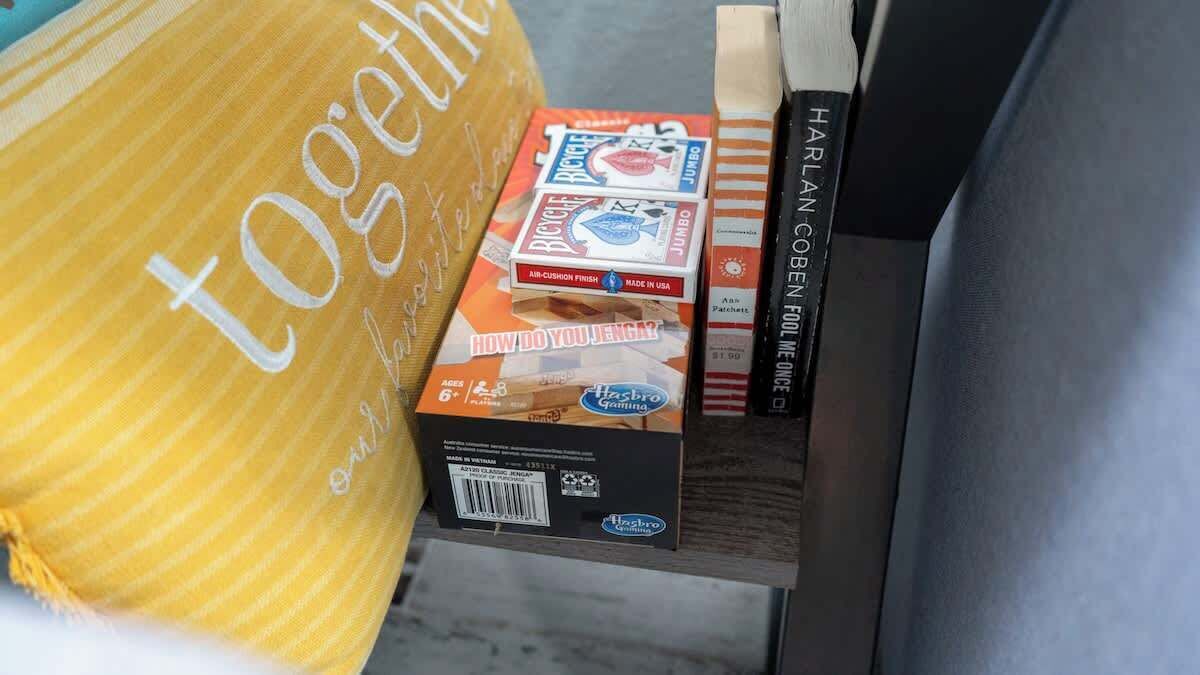 Ein Regal mit einem gestreiften Kissen, einer Kiste mit Hasbro-Puzzle und einem Buch mit dem Titel 'HARLAN COBEN FOOLE'.