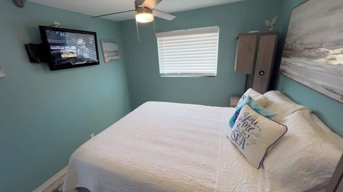 Una camera da letto con un letto, una ventola a soffitto, un televisore, una finestra con persiane e un quadro appeso al muro.
