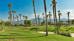 Un campo de golf con palmeras, cielo despejado y montañas a lo lejos.