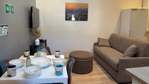 A living room with a sofa, a dining table with bowls and mugs, a mounted TV, and a cityscape painting.