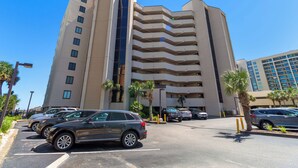 Um hotel de vários andares com design moderno, apresentando um eixo de elevador de vidro central, estacionamento com vários carros e palmeiras.