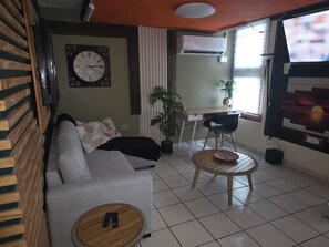 A living room with a sofa, a small round table with remotes, a desk with a chair, a potted plant, and a wall clock.
