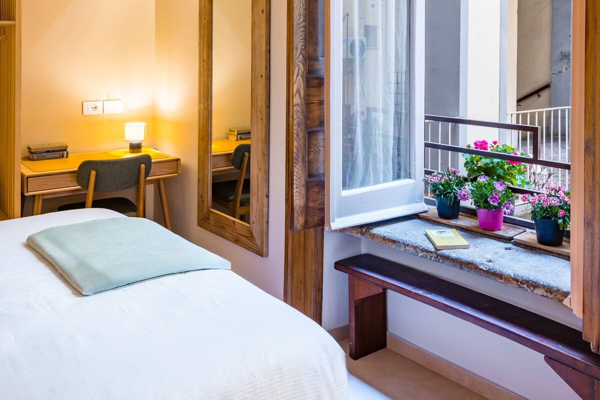 A hotel room with a bed, a desk with a lamp, a large mirror, and a window with potted plants.