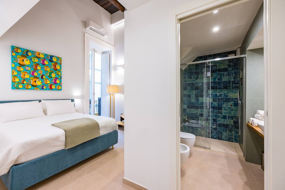 A bedroom with a bed, a colorful fish painting, a bathroom with a glass shower, and a view of the outdoors.