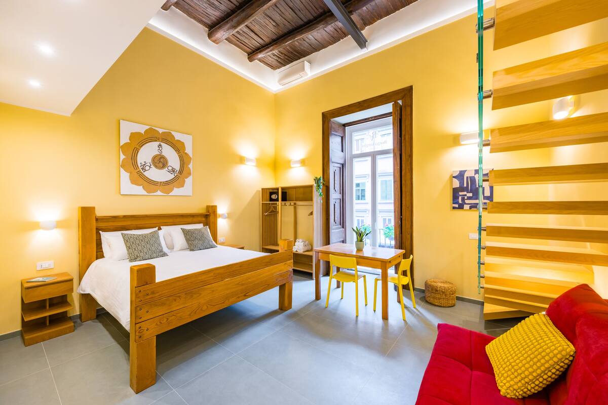 A room with a wooden bed, a red sofa, a small table with chairs, a wall-mounted shelf, and a window with natural light.