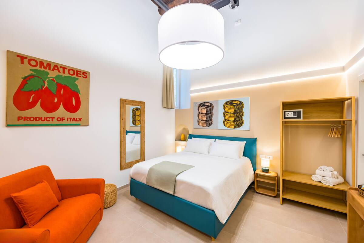 A hotel room with a bed, orange sofa, wooden wardrobe, and a wall with a tomato product sign.