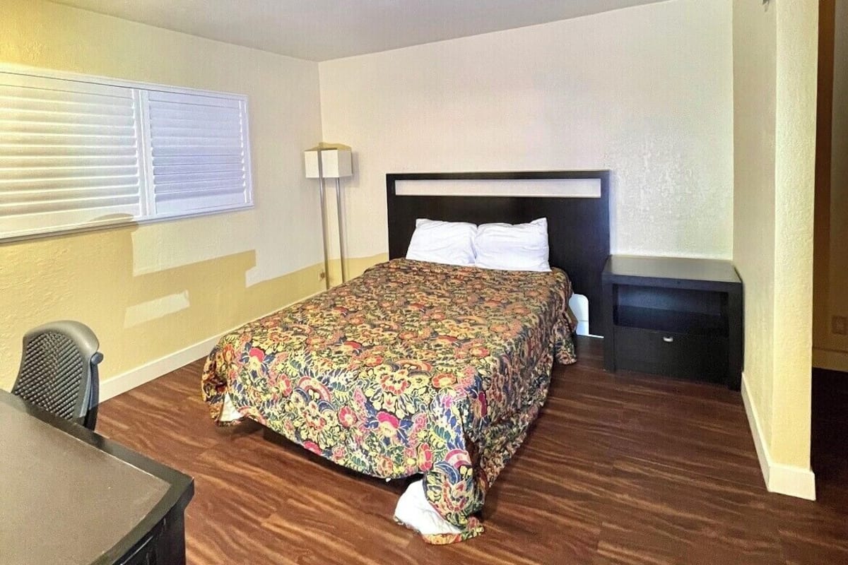 Une chambre d’hôtel avec un lit, un bureau, une chaise, une lampe et une fenêtre.