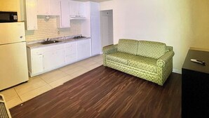 A room with a kitchenette, a green patterned sofa, a bed with a quilt, a desk with a chair, and a red door. - Cloud 9 Motel Cypress Buena Park (Cypress)
