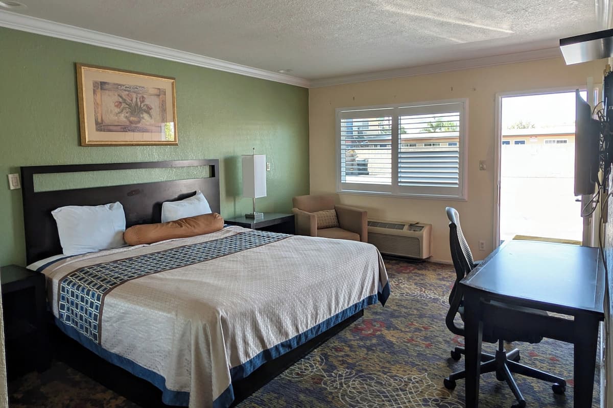 Une chambre d’hôtel avec un lit, un bureau, une chaise, une fenêtre avec des stores et un tableau au mur.