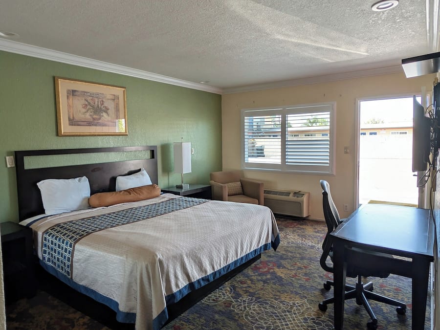 A hotel room with a bed, a desk, a chair, a window with blinds, and a picture on the wall.