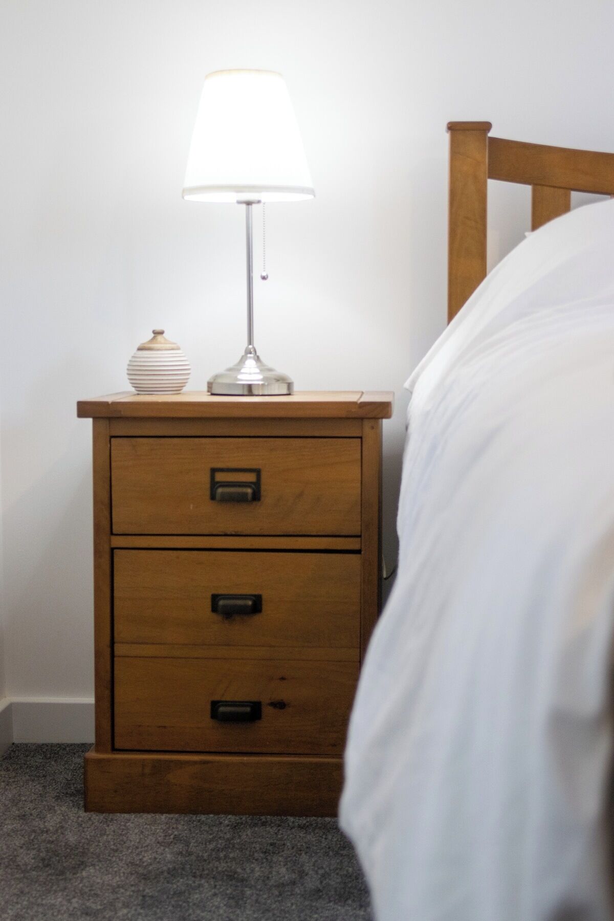 A wooden bedside table with a lamp and a small vase, next to a bed with white bedding.