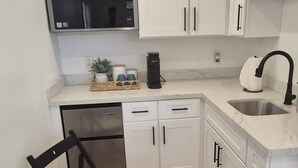 Una cocina compacta con gabinetes blancos, microondas integrado, lavavajillas y un fregadero. Hay una pequeña mesa de comedor con sillas.