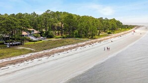 Ein Strand mit einer langen Sandfläche, ein paar Spaziergänger und eine Reihe von Häusern mit einem Wald im Hintergrund.