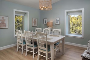 A dining area with a table and chairs, framed artwork on the walls, and windows with a view of trees.