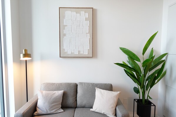 A neutral-toned room with a grey sofa, a framed abstract artwork, a floor lamp, and a potted plant.