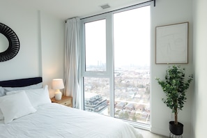 Une chambre avec une grande fenêtre, un lit recouvert de draps blancs, une table de chevet avec une lampe, une plante en pot et un tableau encadré au mur.