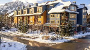 A multi-story hotel with a snow-covered roof, a mountain backdrop, and a clear sky. - Lady Mac Hydeaway at the Crossing (Canmore)