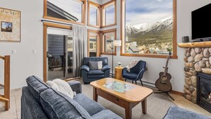 A cozy living room with a fireplace, large windows, and a view of mountains. - Lady Mac Hydeaway at the Crossing (Canmore)