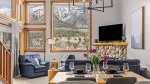A modern living room with a large window, a fireplace, a sofa, a dining table set for two, and a flat-screen TV mounted on the wall. - Lady Mac Hydeaway at the Crossing (Canmore)