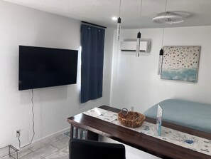 Une pièce dotée d’un téléviseur à écran plat fixé au mur, d’un ventilateur de plafond, d’un appareil de climatisation, d’un lit recouvert d’une couverture bleue, d’une table en bois avec un panier, et d’un tableau encadré accroché au mur.