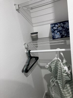 Un coin de pièce avec un mur blanc à la texture particulière, une étagère en métal sur laquelle se trouvent une tasse et un ordinateur portable, et un vêtement rayé qui pend.