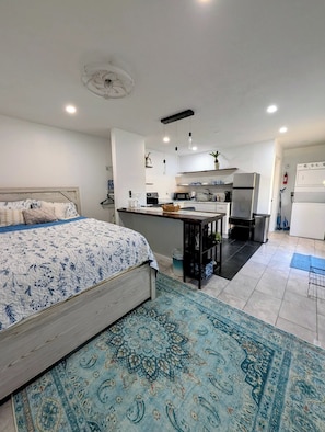 Un dormitorio moderno con una cama grande, una zona de cocina con microondas, refrigerador y lavaplatos, y una pequeña mesa de comedor.