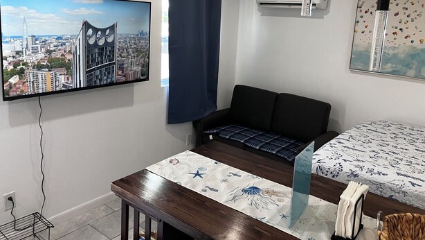 Une pièce avec un téléviseur diffusant une vue sur la ville, un canapé, une table recouverte d’une nappe à motifs et un appareil de climatisation.
