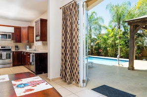 A modern kitchen with stainless steel appliances, a dining area with a table, and a view of a swimming pool and tropical vegetation.