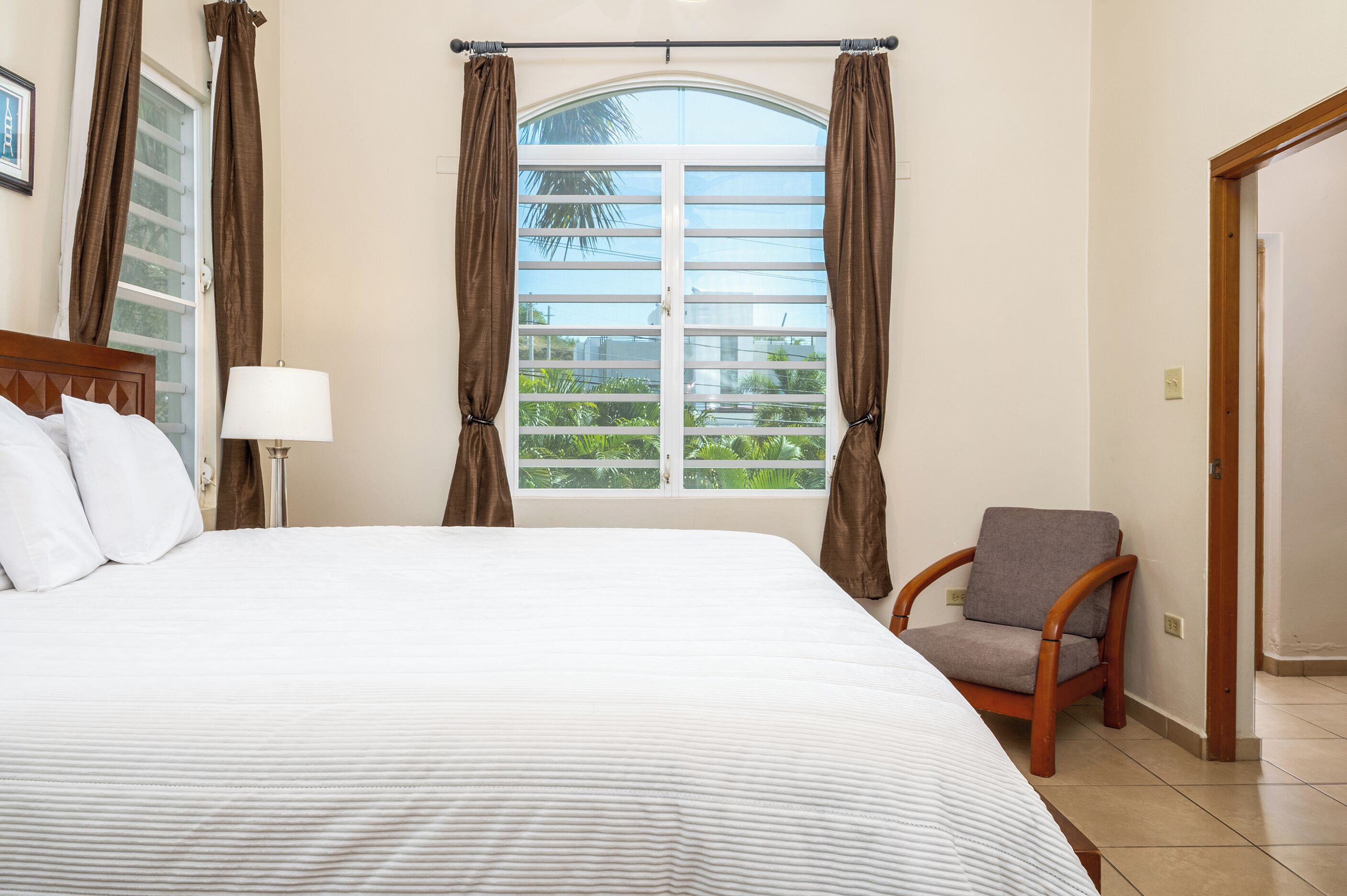 A hotel room with a large bed, a chair, a window with curtains, and a view of greenery outside.