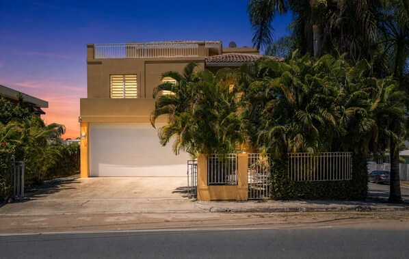 A two-story house with a garage, surrounded by palm trees and a gated entrance.