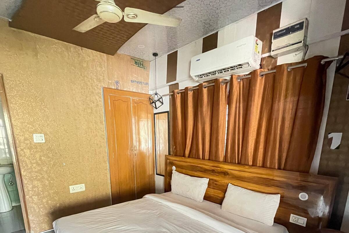 A hotel room with a bed, wooden headboard, brown curtains, an air conditioning unit, and a ceiling fan.