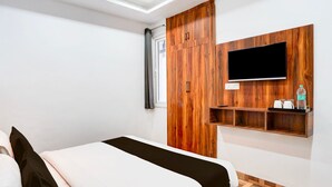 A bedroom with a bed, a wooden wardrobe, a mounted TV, and a shelf with items. - Hotel Elevate Nirvana (Rishikesh)