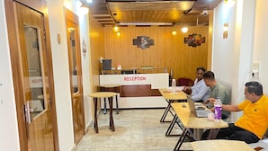 A reception area with a wooden reception desk, tiled flooring, and a corridor leading to a seating area with tables and chairs. - Hotel Elevate Nirvana (Rishikesh)