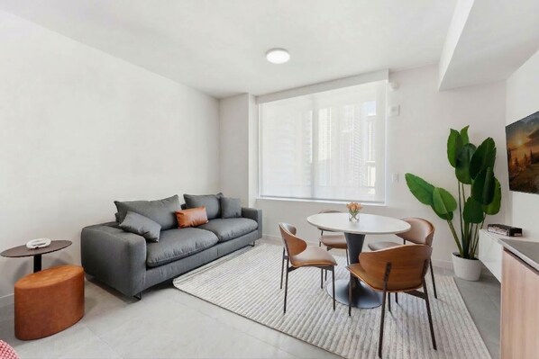 A modern living room with a grey sofa, a round dining table with chairs, a small side table, a large potted plant, and a window with a city view. - Miami 176 - One Bedroom Apartment, Sleeps 3 (Miami)