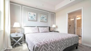 Une chambre à coucher comprenant un lit, deux tables de chevet, une commode, une porte donnant sur une salle de bain et deux tableaux accrochés au mur.