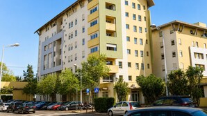 Un edificio de varios pisos con fachada curva, múltiples balcones y un estacionamiento con coches y un sendero peatonal.