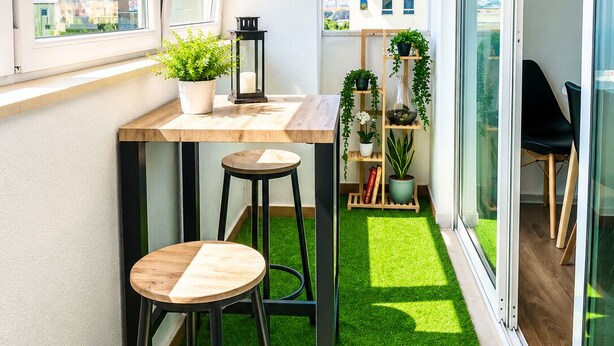 Un balcón con mesa de madera, dos taburetes, una planta en maceta y una estantería.