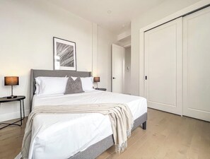 Un lit bien fait, avec du linge de lit blanc, une tête de lit grise et un seul oreiller. Un tableau encadré est accroché au mur au-dessus du lit, et une porte fermée se trouve à droite.