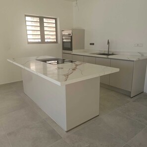A kitchen with a central island, a built-in oven, and a window with blinds. - Luxury apartment, including pool, 6 minutes from Mambo Beach. (Willemstad)