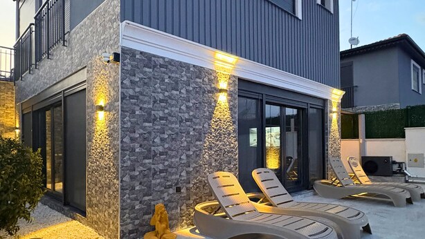 Una casa moderna con fachada de piedra, un balcón y una zona de piscina con sillones de descanso.