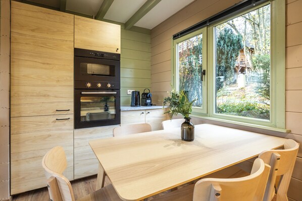 A kitchen with wooden cabinets, a built-in oven, a dining table with chairs, and a window with a view of greenery. - The Renardeau cabin for 5 people is a cozy, fully renovated accommodation featuring two bedrooms, a modern shower room, a premium kitchen, and a living area with a pellet stove. (Dochamps)