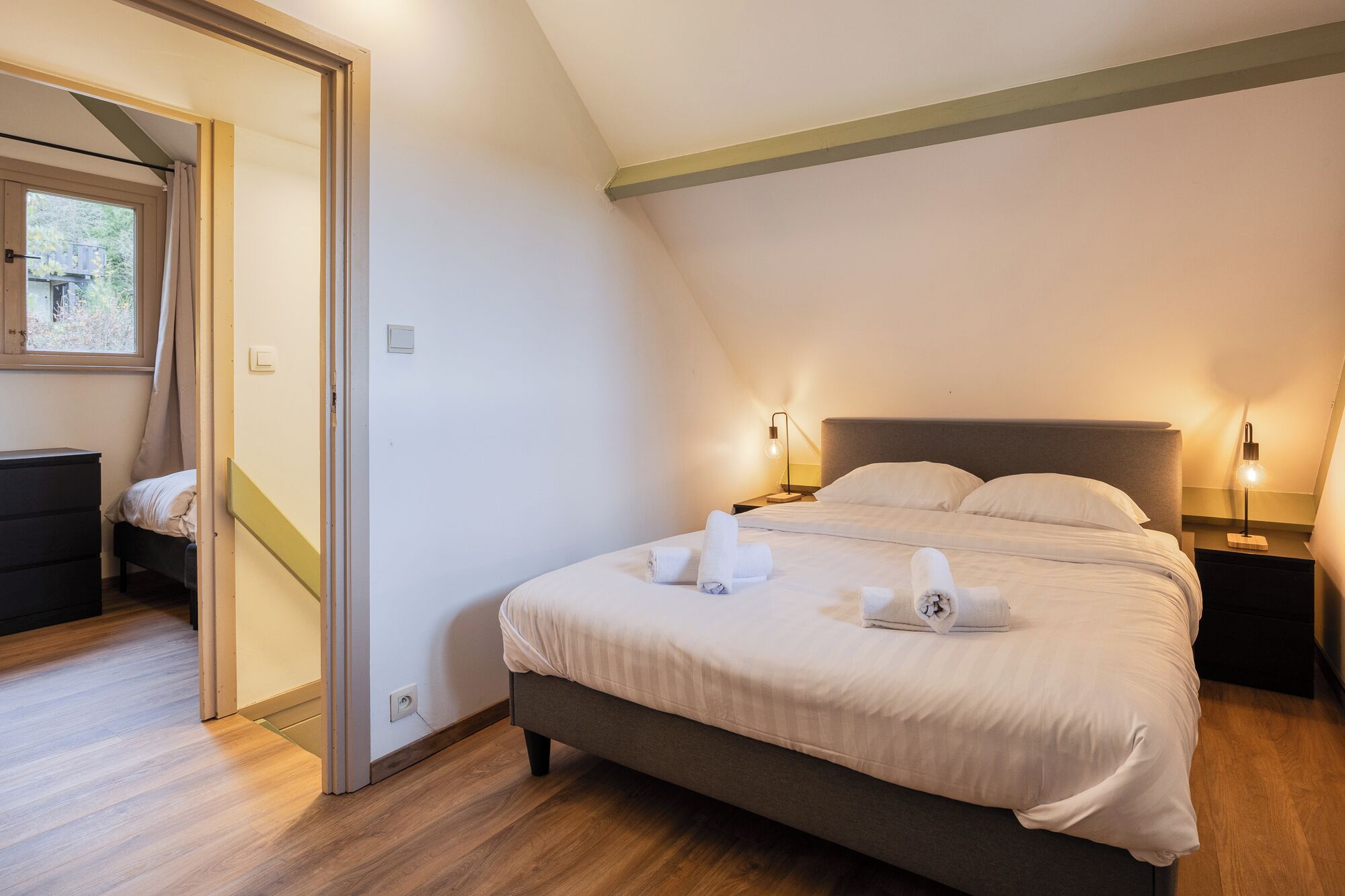 A bedroom with a bed, bedside lamps, a nightstand, a dresser, and a door leading to another room.