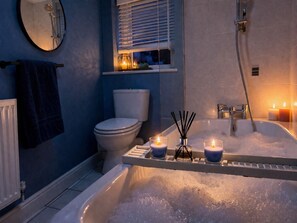 A bathroom with a bathtub filled with bubbles, a toilet, a window with blinds, and candles on the windowsill.