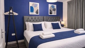 Quarto de hotel com uma cama grande, cabeceira estofada cinza, dois quadros abstratos na parede, um cabideiro suspenso e um abajur.