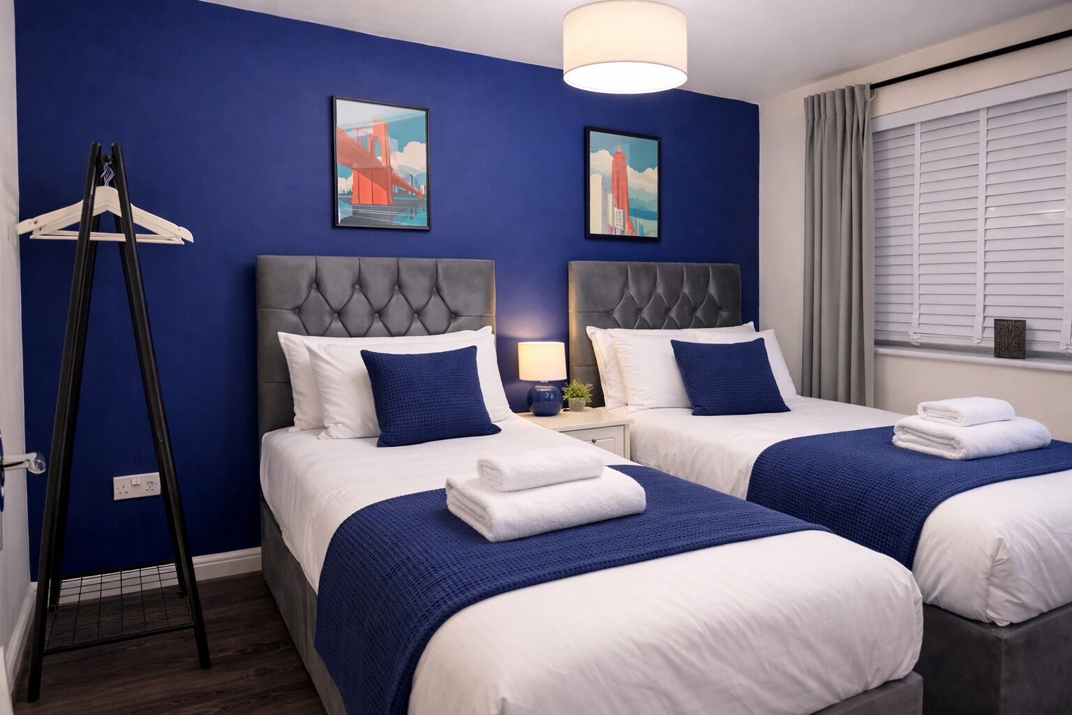 Ein Hotelzimmer mit zwei Betten, einem grauen, gesteppten Kopfteil, weißer Bettwäsche, blauen Zudecken und Bildern an den Wänden.