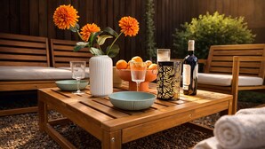 Uma mesa de madeira com um vaso de flores laranjas, uma tigela de laranjas, uma garrafa de vinho e um candeeiro com padrão.