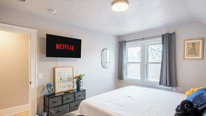 Makuuhuoneessa on sänky, lipasto, seinälle asennettu televisio, ikkuna, jossa on verhot, ja seinällä oleva kehystetty kuva.